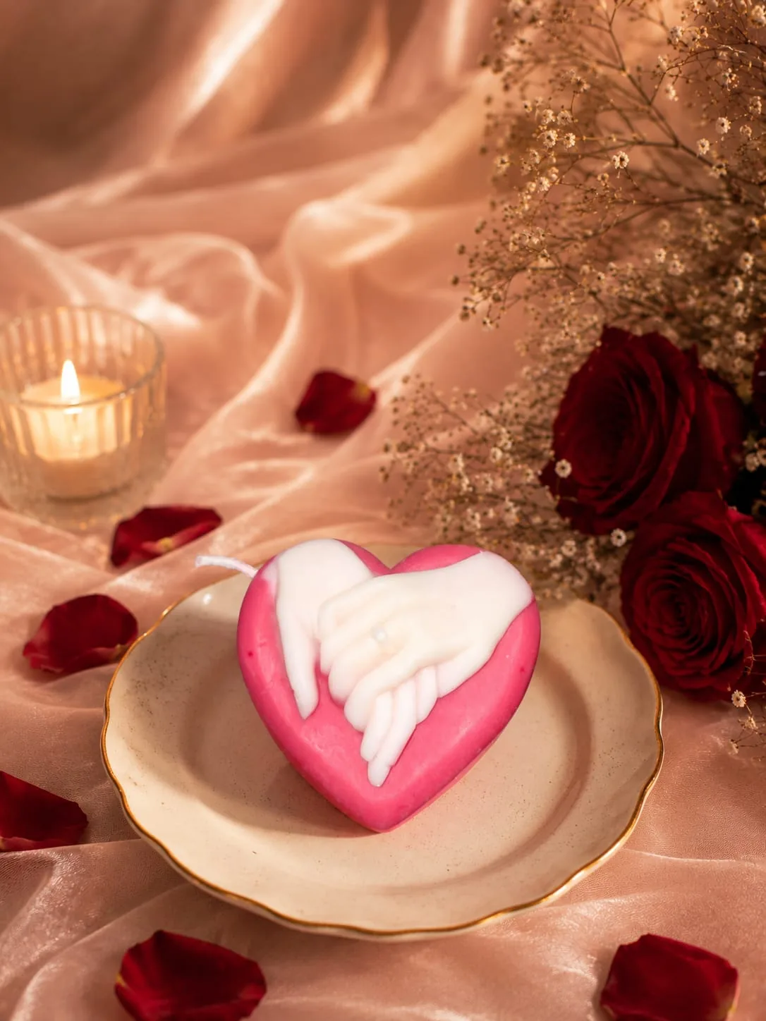 Pink and white heart candle with clasped hands, on a dish surrounded by rose petals